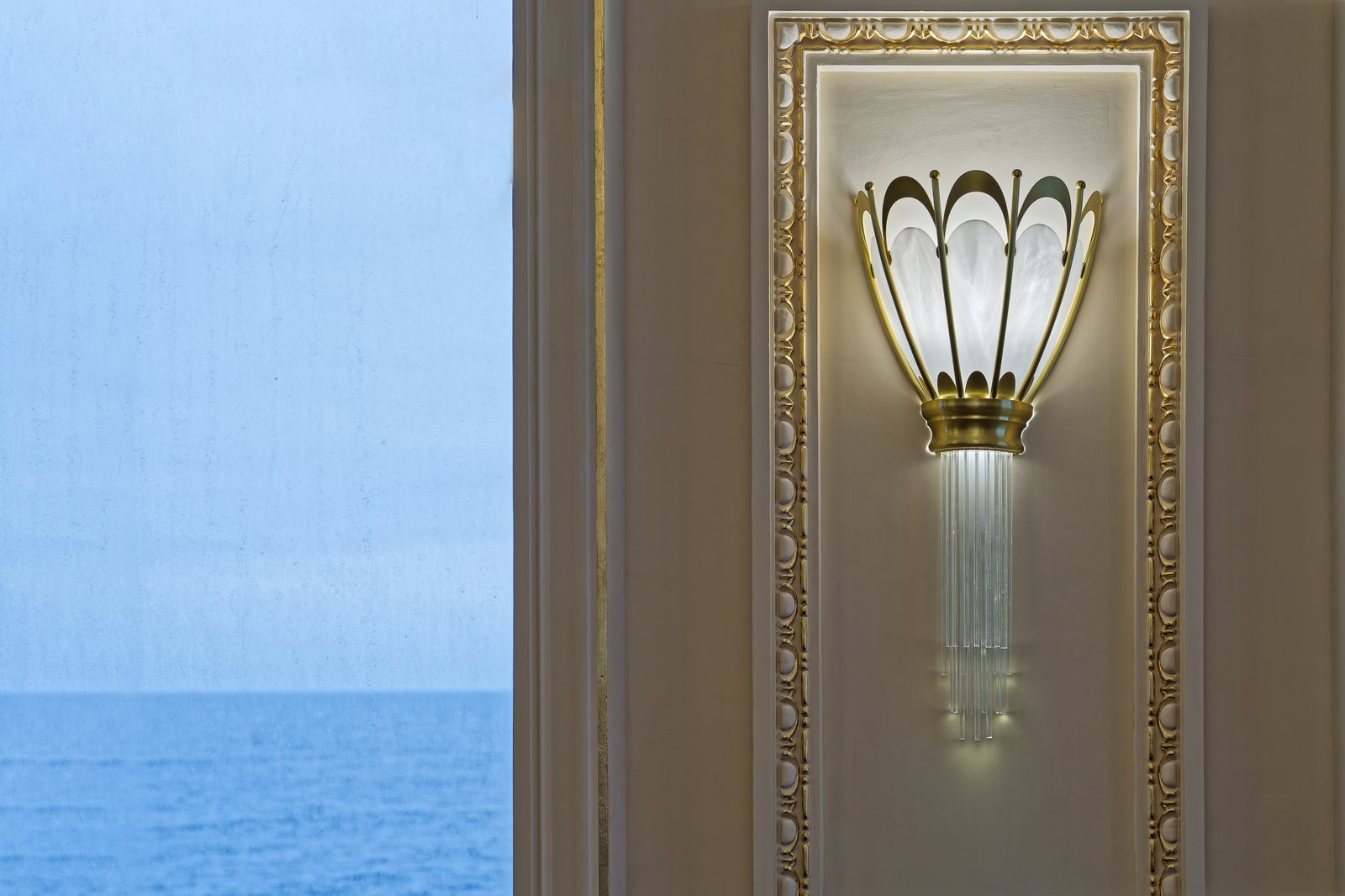 Aplică Art Nouveau aprinsă pe un perete cu profile aurite, lângă o fereastră cu vedere la mare