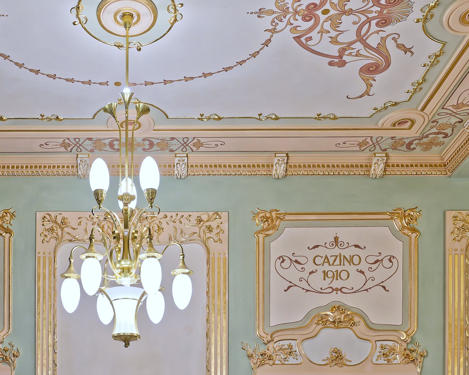 Candelabru de alamă cu brațe și globuri de sticlă opal, sub plafonul pictat cu inscripția „Cazino 1910”