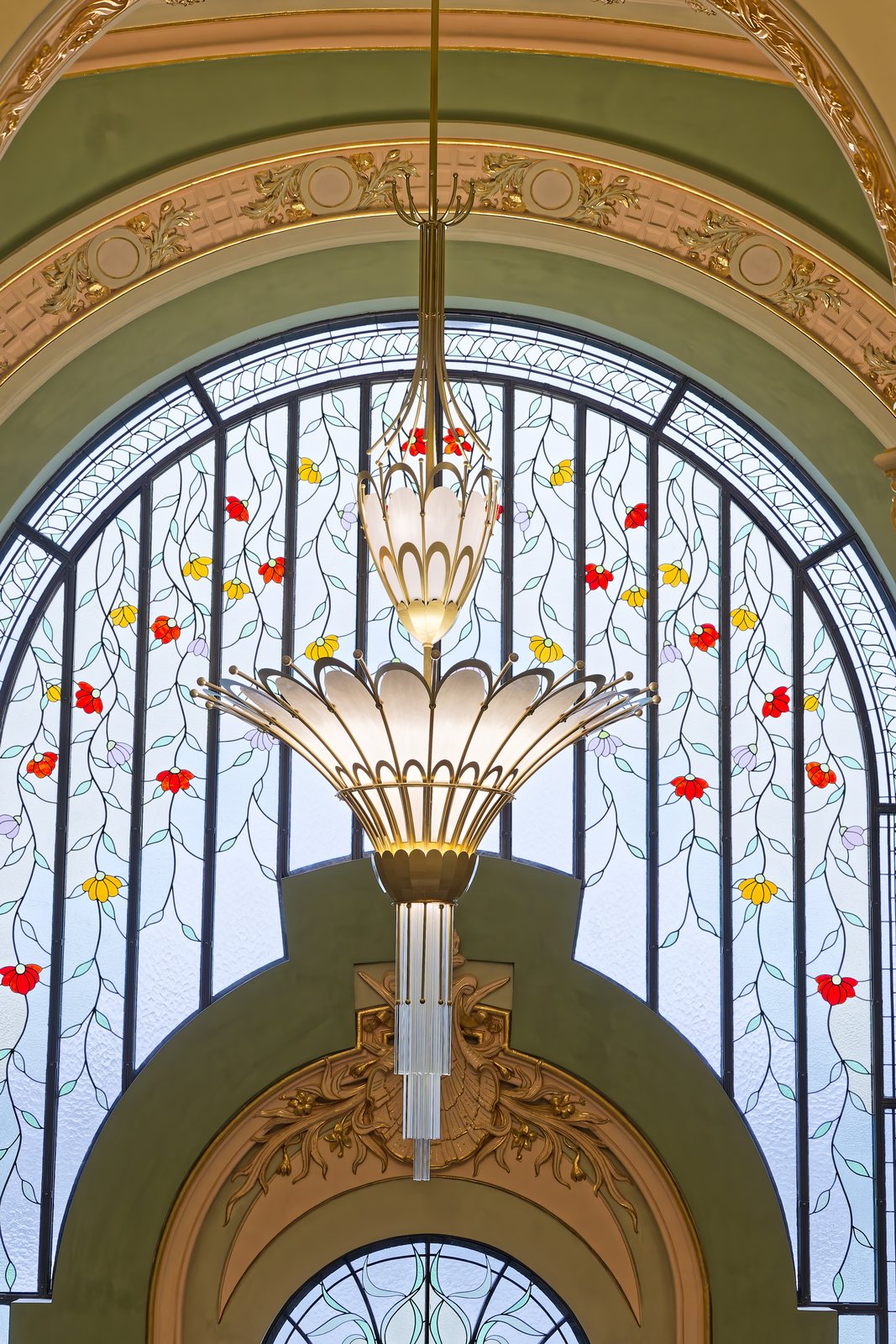 Candelabru Art Nouveau suspendat în fața unui vitraliu mare cu motive florale, pe scara de onoare