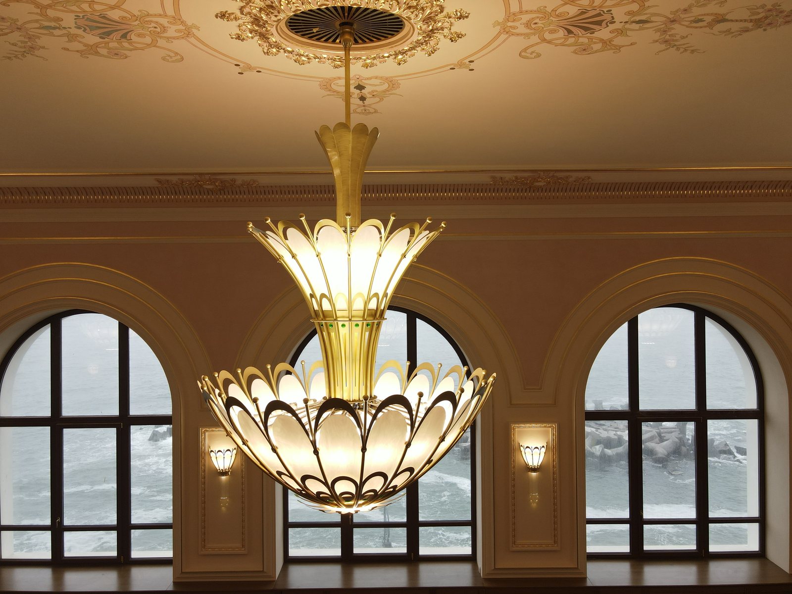 Candelabru Art Nouveau din alamă și sticlă opal, fotografiat de la nivelul lui, cu marea vizibilă prin ferestrele arcuite